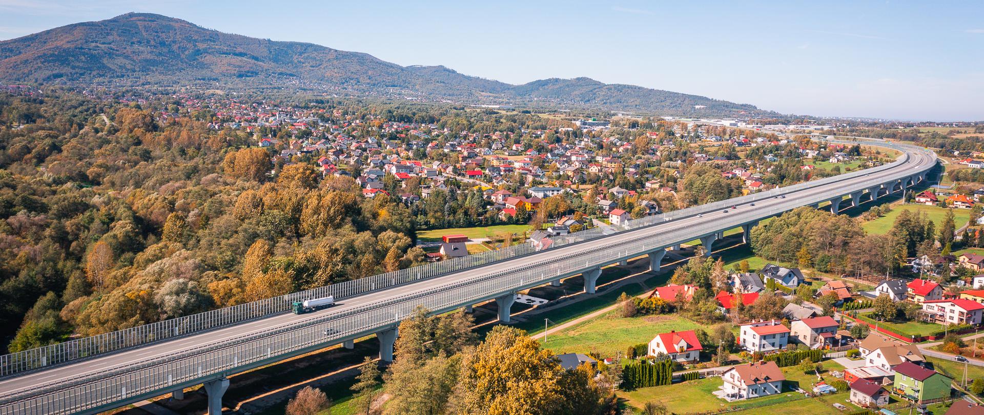 Na zdjęciu przedstawiono szeroką panoramę górzystego terenu, na którym rozciąga się nowoczesny wiadukt drogowy. Droga biegnie wysoko ponad terenem zabudowanym, prowadząc przez tereny wiejskie z małymi, kolorowymi domami o czerwonych dachach. W tle widoczne są zalesione wzgórza, tworzące malowniczy krajobraz. Wiadukt łączy różne części miejscowości, przecinając doliny i małe rzeki, a na jego powierzchni można dostrzec pojazdy. Fotografię wykonano prawdopodobnie jesienią, ponieważ drzewa są częściowo pokryte złotymi i brązowymi liśćmi.