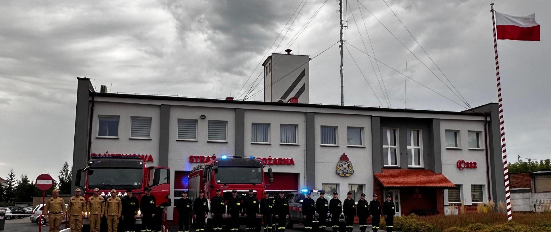 Na zdjęciu obsada jednostki ratowniczo - gaśniczej oraz druhowie OSP oddający hołd zmarłym kolegom z Poznania. Strażacy ustawieni w szyku przed obiektem komendy na tle pojazdów pożarniczych z włączonymi sygnałami świetlnymi