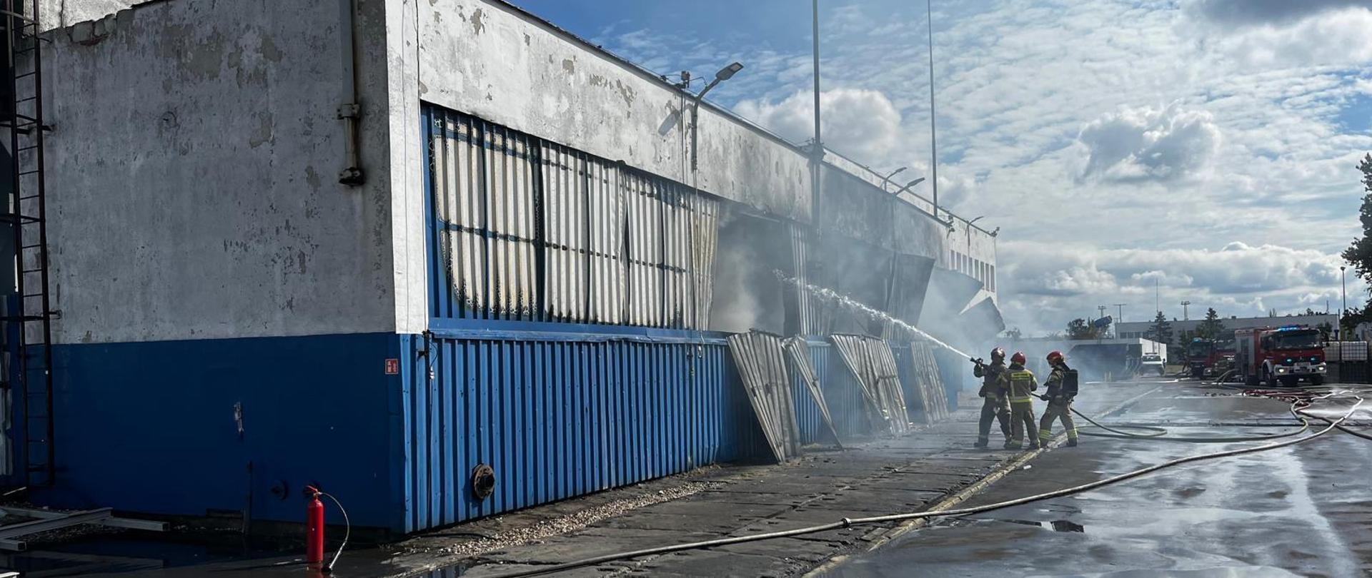 Grupa strażaków strażaków gaszących pożar magazynu technicznego. Z budynku wydobywa się dym. 