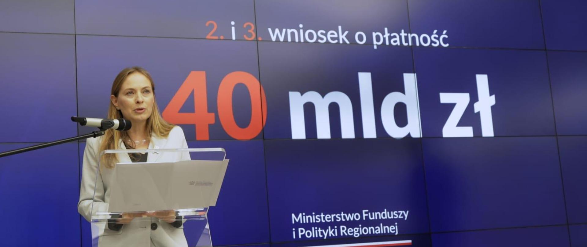 Minister Katarzyna Pełczyńska-Nałęcz stoi z lewej strony przy szklanym pulpicie i mówi do mikrofonu.