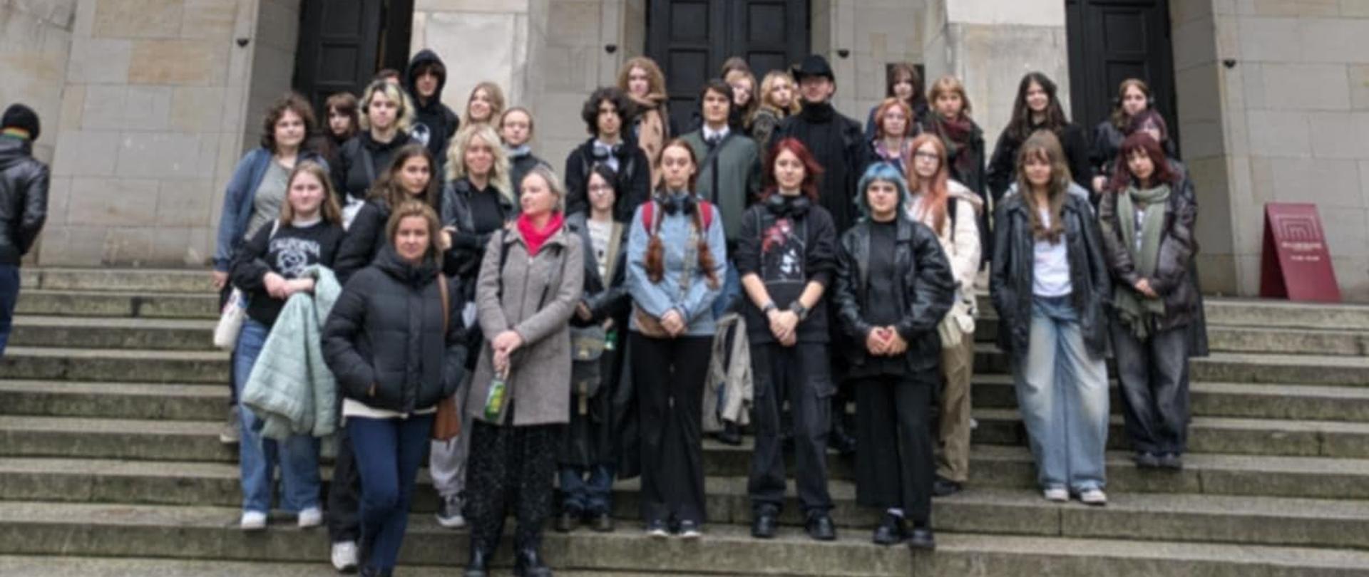 Zdjęcie grupowe, pozowane. Uczniowie i nauczycielki, Aneta Lekan, Paulina Zarębska stoją przed wejściem do Muzeum Narodowego w Warszawie. Na jego frontowej ścianie znajduje się wielki, trzyczęściowy baner reklamujący wystawę obrazów J. Chełmońskiego od 27.09 do 26.01. Na banerze oprócz dat, napisu Józef Chełmoński reprodukcja pejzażu artysty ukazująca ptaka w locie nisko akwenu i roślinności.