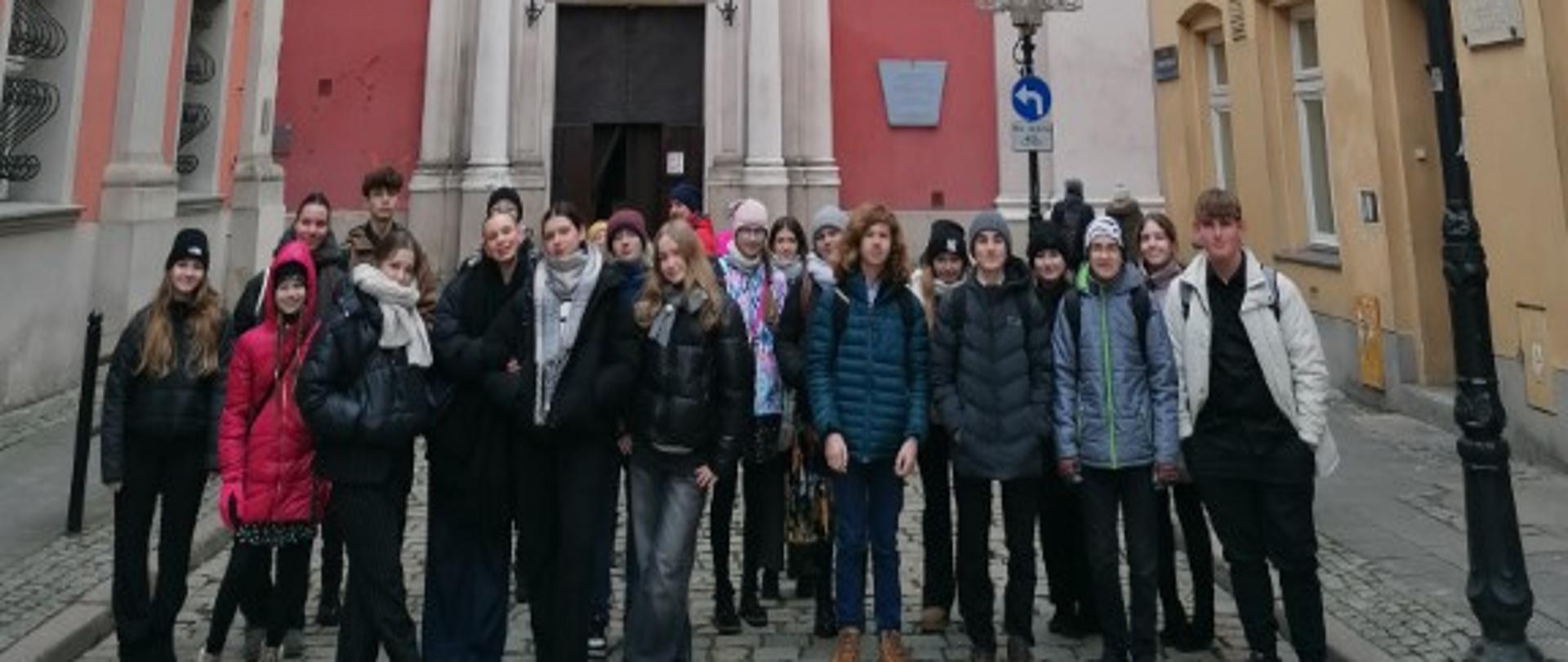 Zdjęcie kolorowe. Grupa uczniów przed fasadą barokowego kościoła.