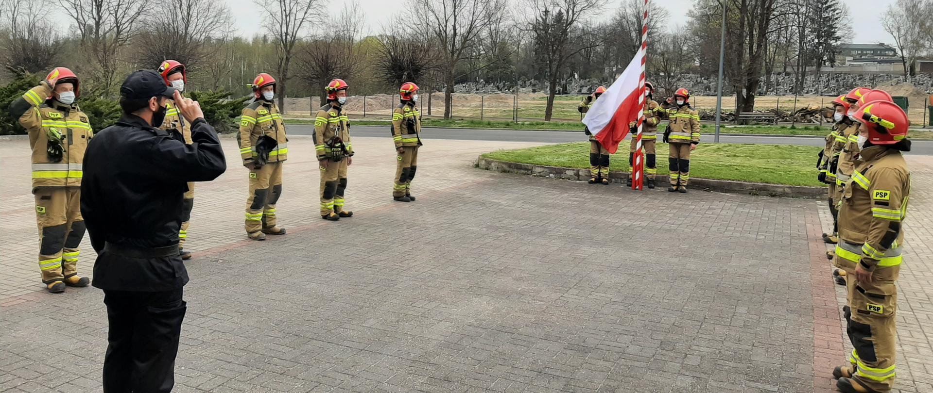 Na zdjęciu widać 14-stu strażaków. 10-ciu z nich stoi w dwóch 5-cio osobowych szeregach, zwróconych twarzami do siebie. Między nimi znajduje się strażak (dowódca) zwrócony twarzą do masztu z flagą znajdującym się na przeciw niego. Flagę polski na maszt wciąga 3 strażaków.
