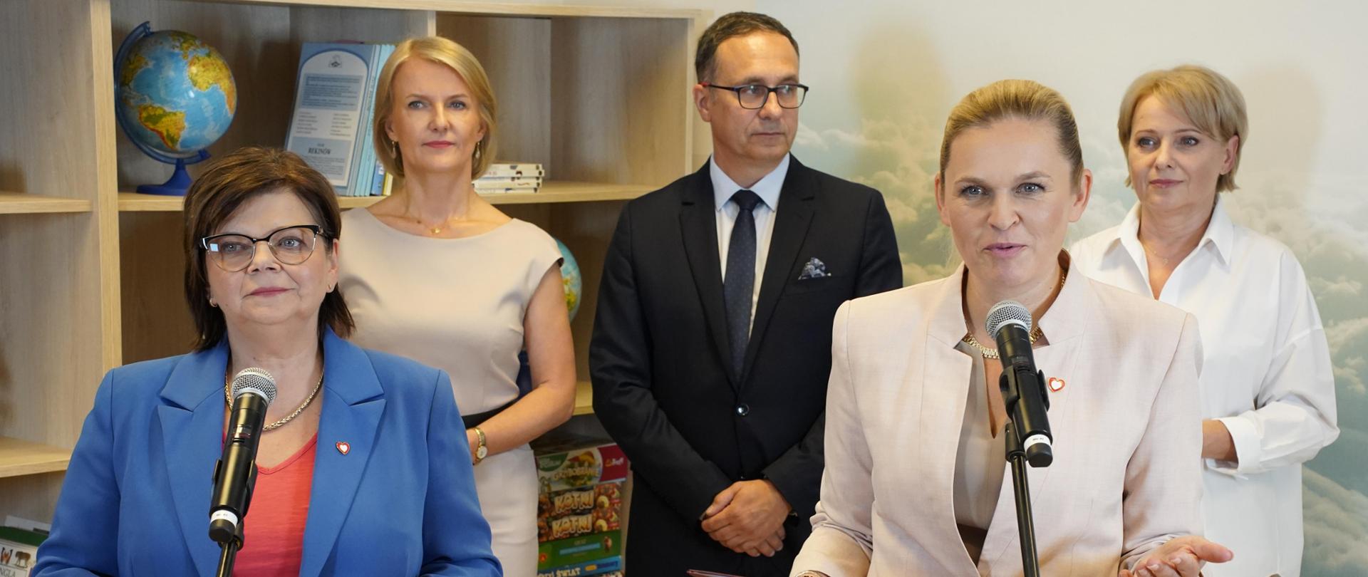 Na pierwszym od lewej Minister Zdrowia Izabela Leszczyna oraz Minister Edukacji Barbara Nowacka stoją przy mikrofonach.
Za nimi znajdują się trzy osoby. Kobieta ubrana w białą sukienkę, mężczyzna w środku ubrany w ciemny garnitur z krawatem i kobieta ubrana w biały strój. W tle półka na książki z globusem i książkami są zauważalnymi elementami.