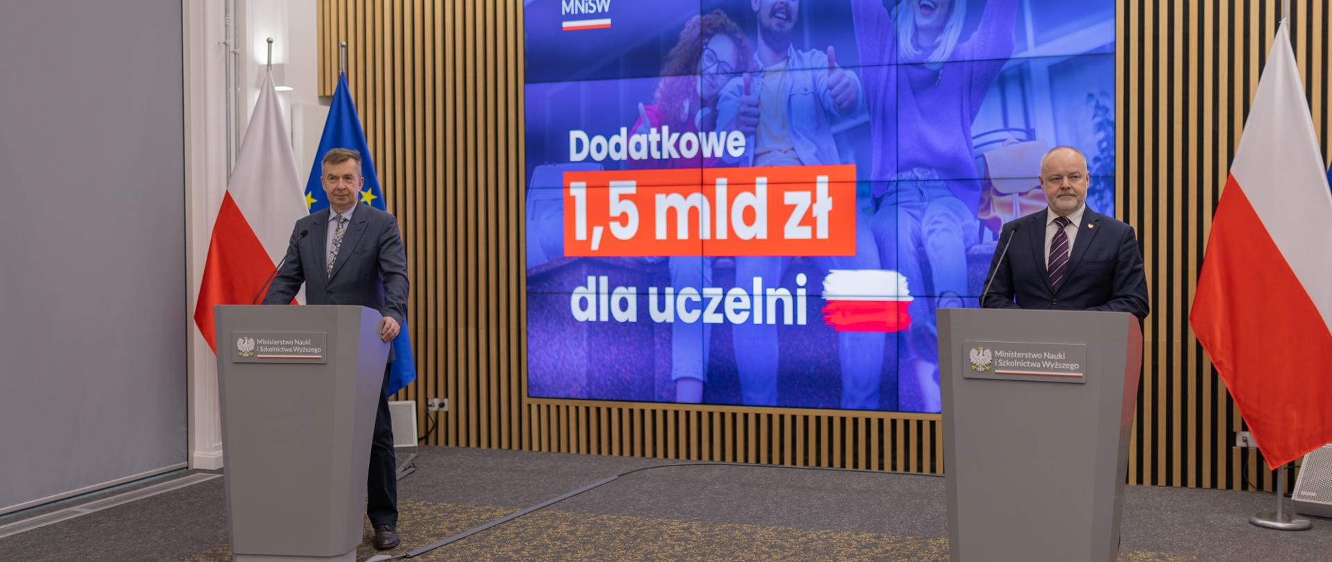 Za dwiema mównicami stoją minister Wieczorek i wiceminister Gzik, za nimi wbudowany w ścianę wielki niebieski ekran z napisem Dodatkowe 1,5 mld zł dla uczelni.