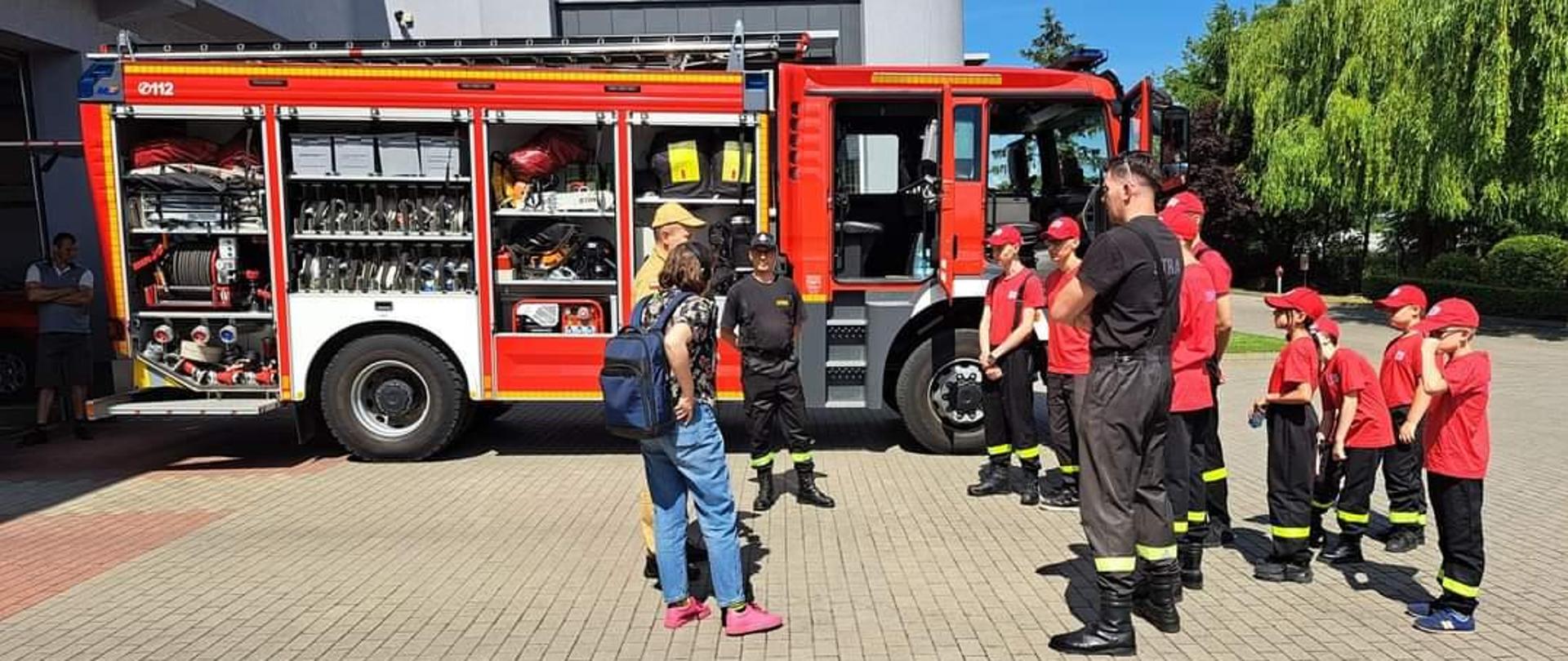 Zdjęcie przedstawia strażaka, który podczas dnia otwartych strażnic omawia dzieciom zagrożenia jakie mogą wystąpić w domach mieszkalnych oraz opowiada o pracy strażaka.