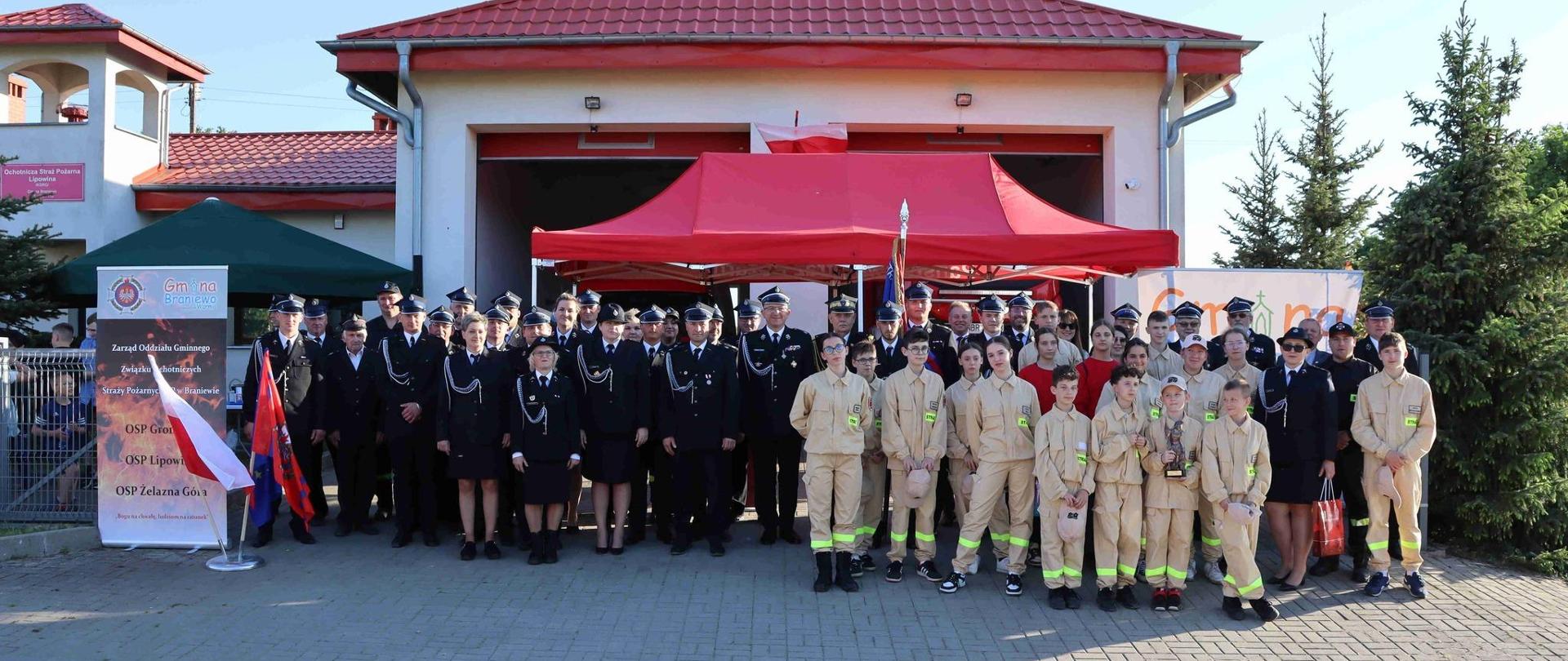 Na tle strażnicy OSP stoją strażacy w mundurach galowych oraz dzieci w jasnych mundurkach.