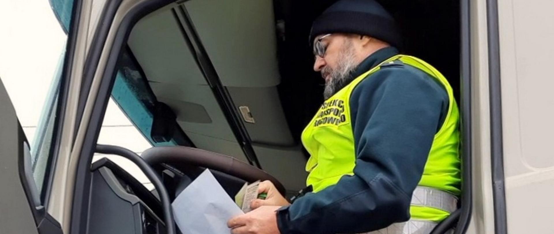 Inspektor siedzi na miejscu kierowcy w kabinie ciężarówki. W dłoni trzyma dokumenty.