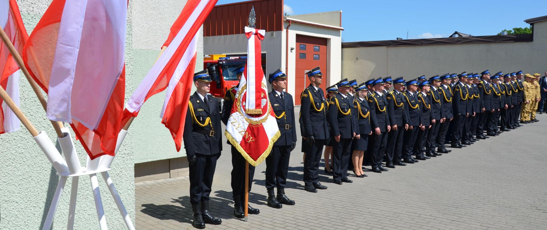 Strażacy Komendy ustawieni w szeregu, galowy ubiór, na pierwszym planie flagi państwowe i poczet sztandarowy
