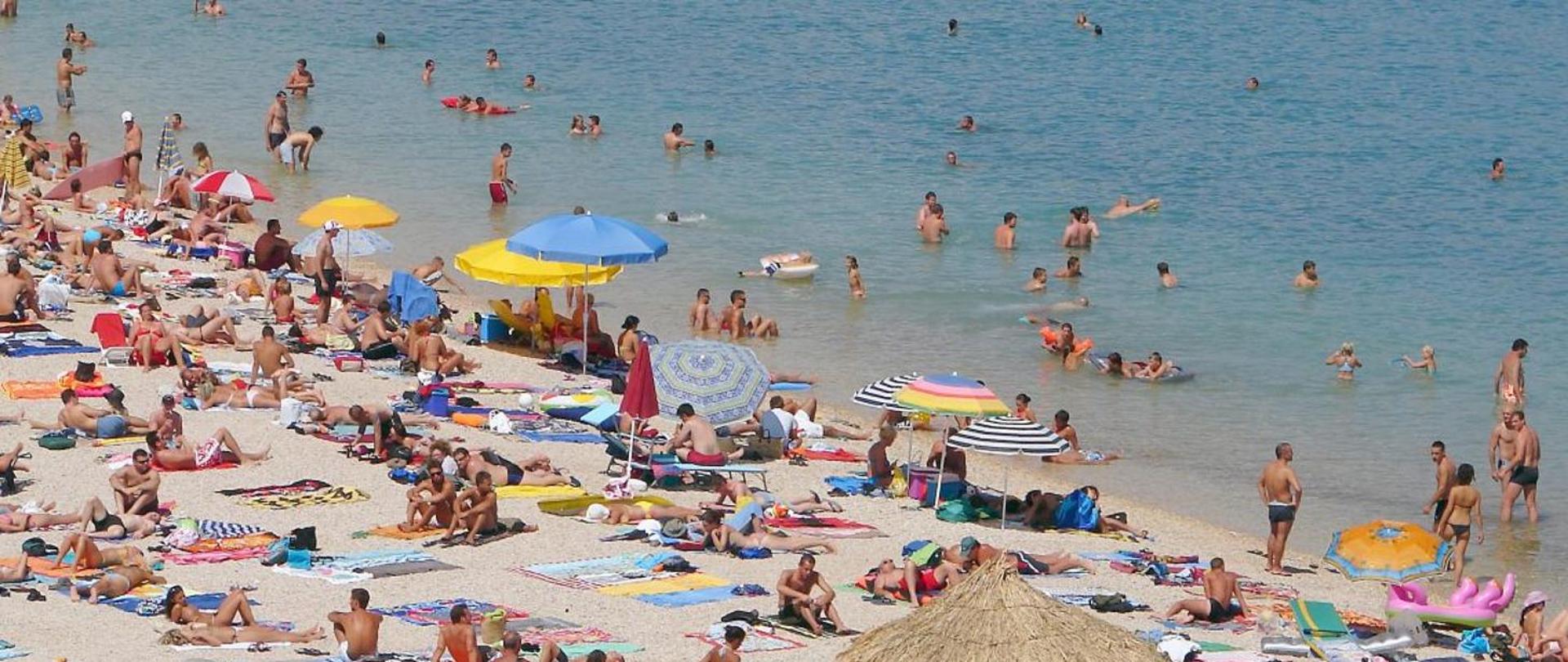 Zdjęcie przedstawia dużą grupę ludzi opalających się na plaży i pływających w jeziorze.