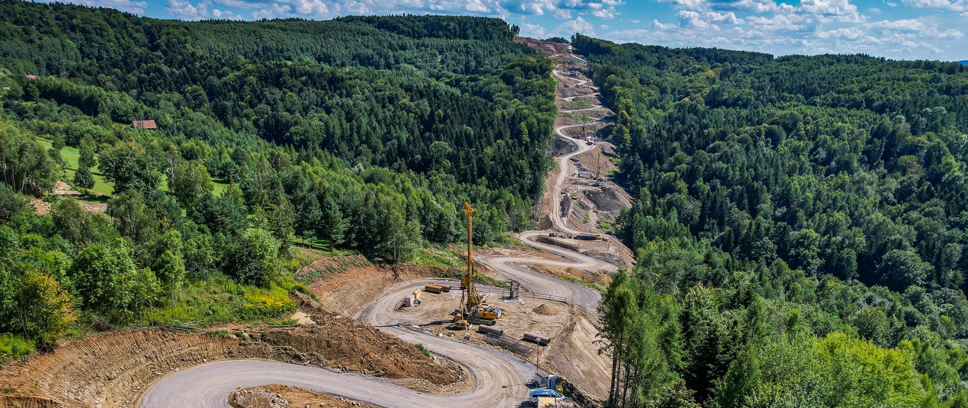 Widok lotniczy na budowę S19 między Babicą a Jawornikiem. Świeci słońce. Widać wzgórze, lasy a po środku drogę technologiczną biegnącą ja serpentyny. U podnóżą palownica. 