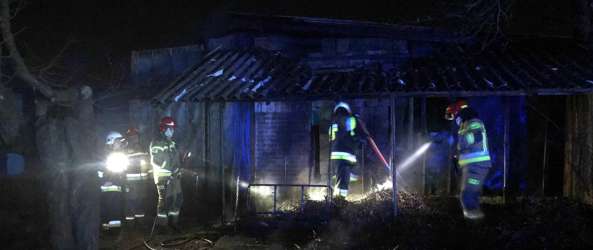 zdjęcie zrobione w porze nocnej i przedstawia pięciu ratowników podczas przerzucania nadpalonego drewna opałowego i przelewania wodą. W tych działań ratowniczo-gaśniczych wykorzystano sprzęt burzący oraz wodę z szybkiego natarcia.