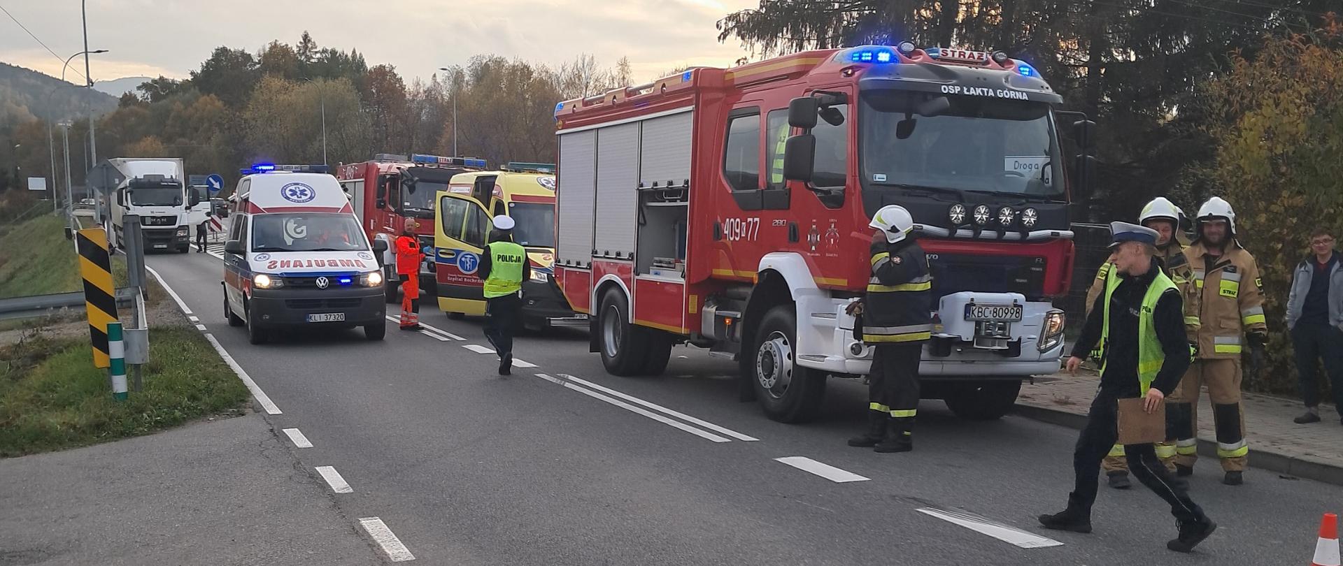 Zdjęcie przedstawia wypadek dwóch samochodów osobowych. Na zdjęciu widać po lewej stronie biały pojazd Zespołu Ratownictwa Medycznego. Po prawej stronie stoi czerwony pojazd pożarniczy a za nim ustawiony jest żółty pojazd ZRM a zanim kolejny czerwony pojazd pożarniczy. Koło karetek stoi ratownik medyczny. Przed pojazdami pożarniczymi widać strażaków ubranych w czarne i piaskowe ubrania specjalne na głowie maja hełmy. Obok nich widać dwóch policjantów prowadzących czynności dochodzeniowe. Po prawej stronie widać na drodze pachołek ostrzegawczy