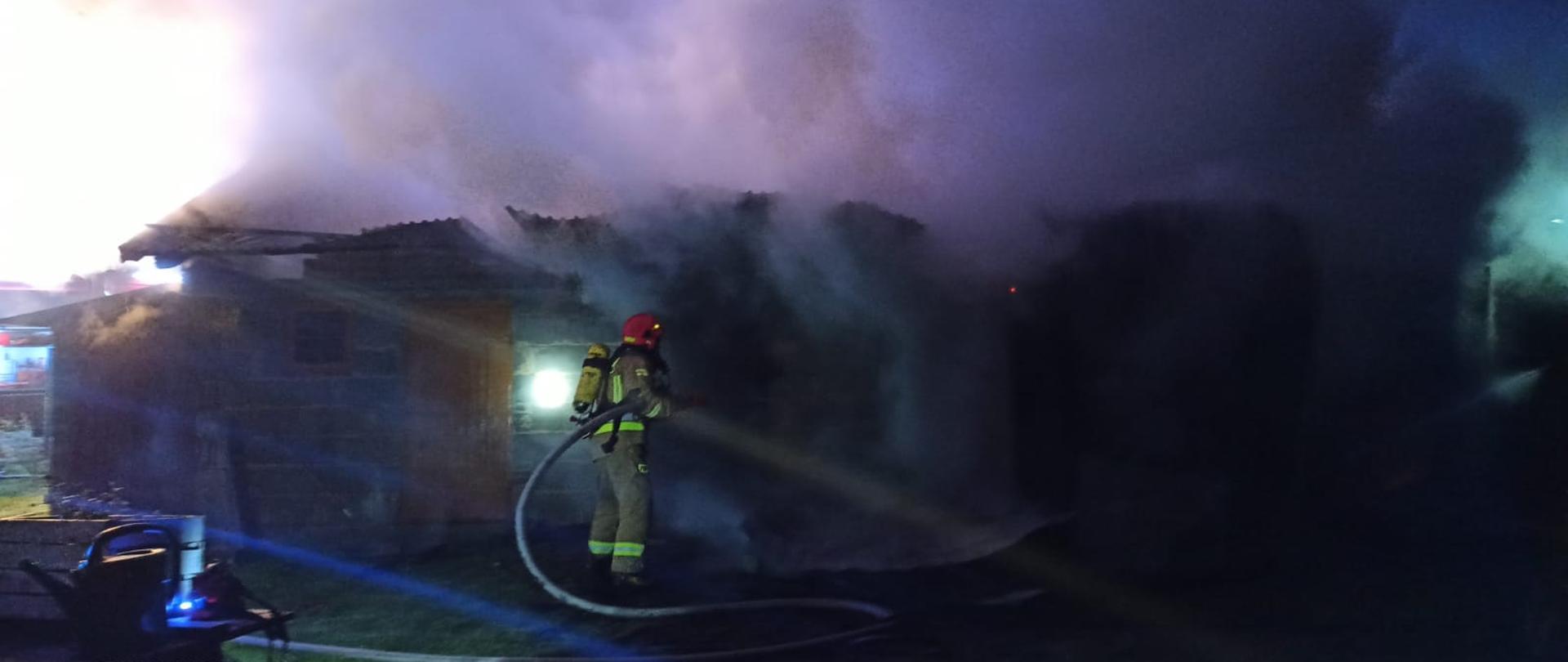 Zdjęcie obrazuje strażaka trzymającego w rękach prądownicę oraz wąż pożarniczy. Strażak w umundurowaniu bojowym z założonym na plecach aparatem ochrony dróg oddechowych. Zdjęcie zrobione w porze nocnej. Przed strażakiem znajduje się murowany budynek gospodarczy spowity gęstym dymem pożarowym. 