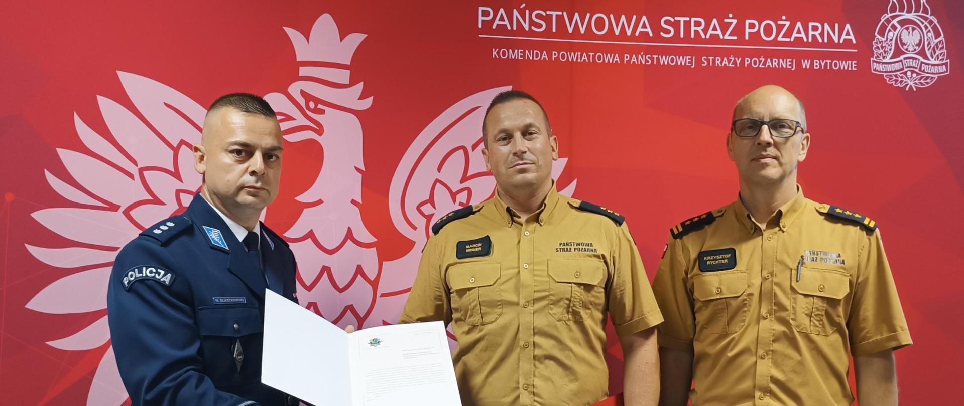 Dwóch funkcjonariuszy straży pożarnej w umundurowaniu koloru piaskowego i funkcjonariusz policji w mundurze koloru granatowego na tle czerwonego baneru Państwowa Straż Pożarna. Funkcjonariusz policji prezentuje list gratulacyjny. 
