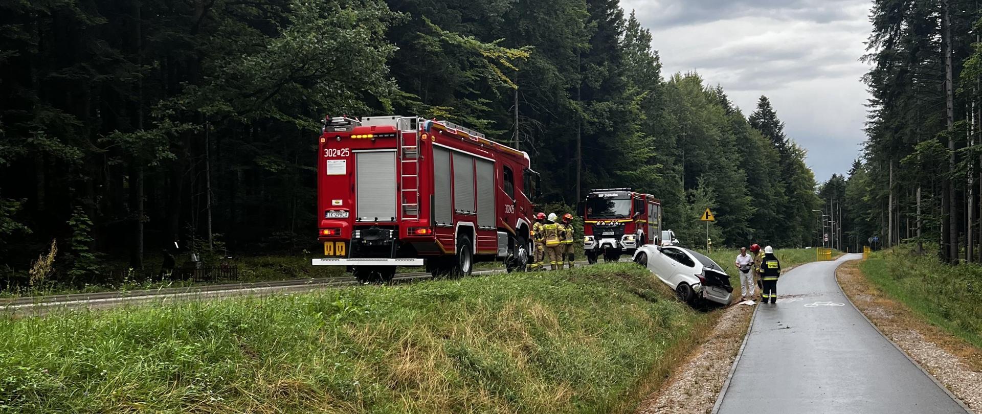 Zdjęcie przedstawia drogę powiatową i ścieżkę rowerową w Siodłach. Na jezdni stoją dwa samochody pożarnicze, a w przydrożnym rowie stoi jeden z uszkodzonych w wyniku kolizji pojazdów. 