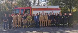 Zdjęcie grupowe na tle pojazdu pożarniczego. Na zdjęciu widać osoby cywilne oraz strażaków ubranych w mundury specjalne, koszarowe oraz służbowe.