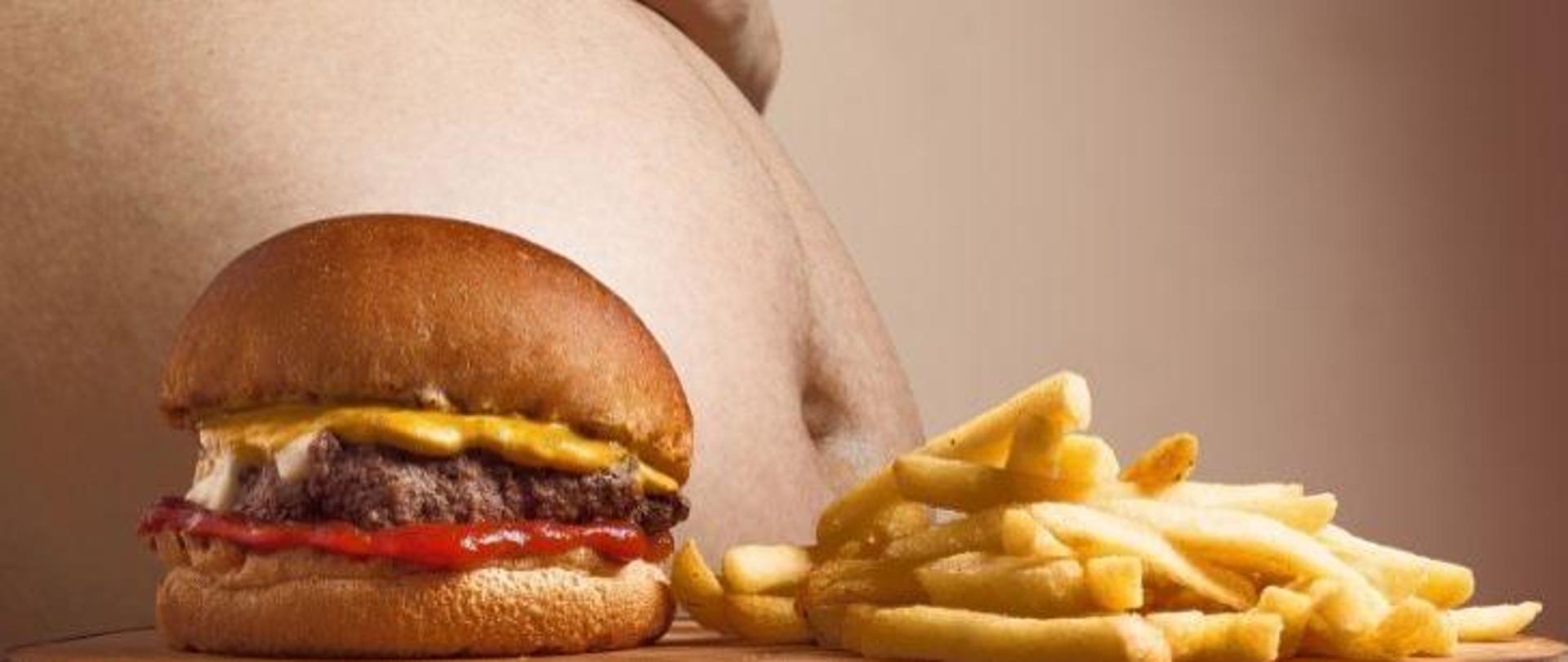 Na pierwszym planie widać tłustego hamburgera wraz z frytkami. Na drugim planie widać brzuch osoby otyłej.