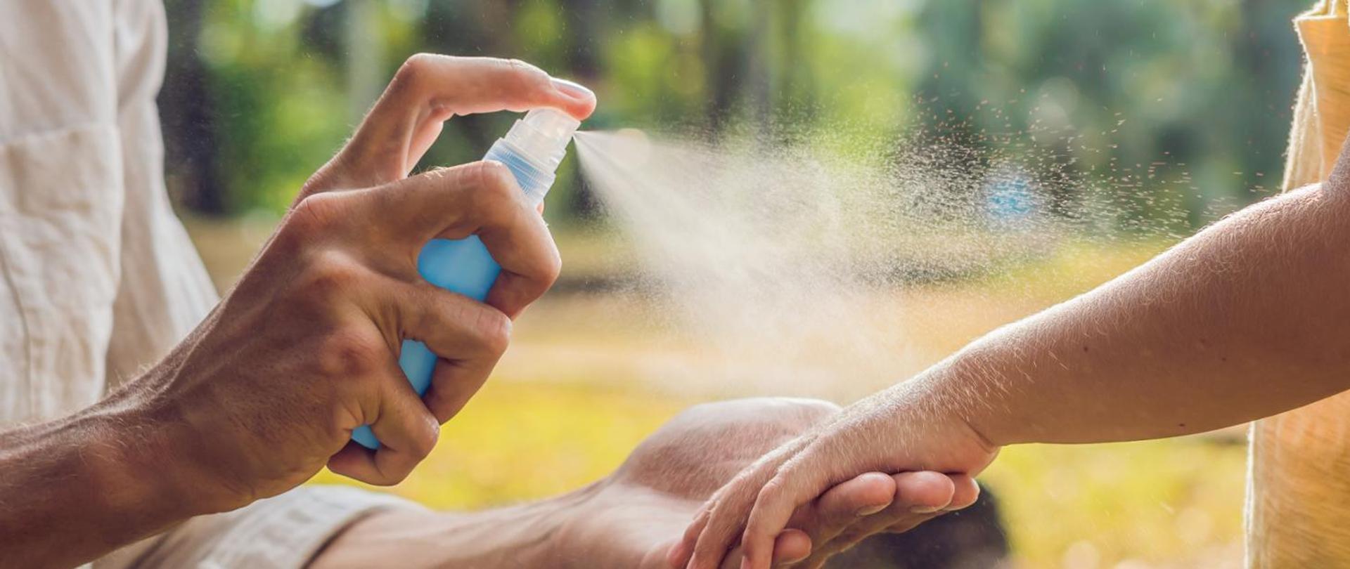 Z lewej ręka trzymająca spray, psikający na rękę dziecka po prawej.