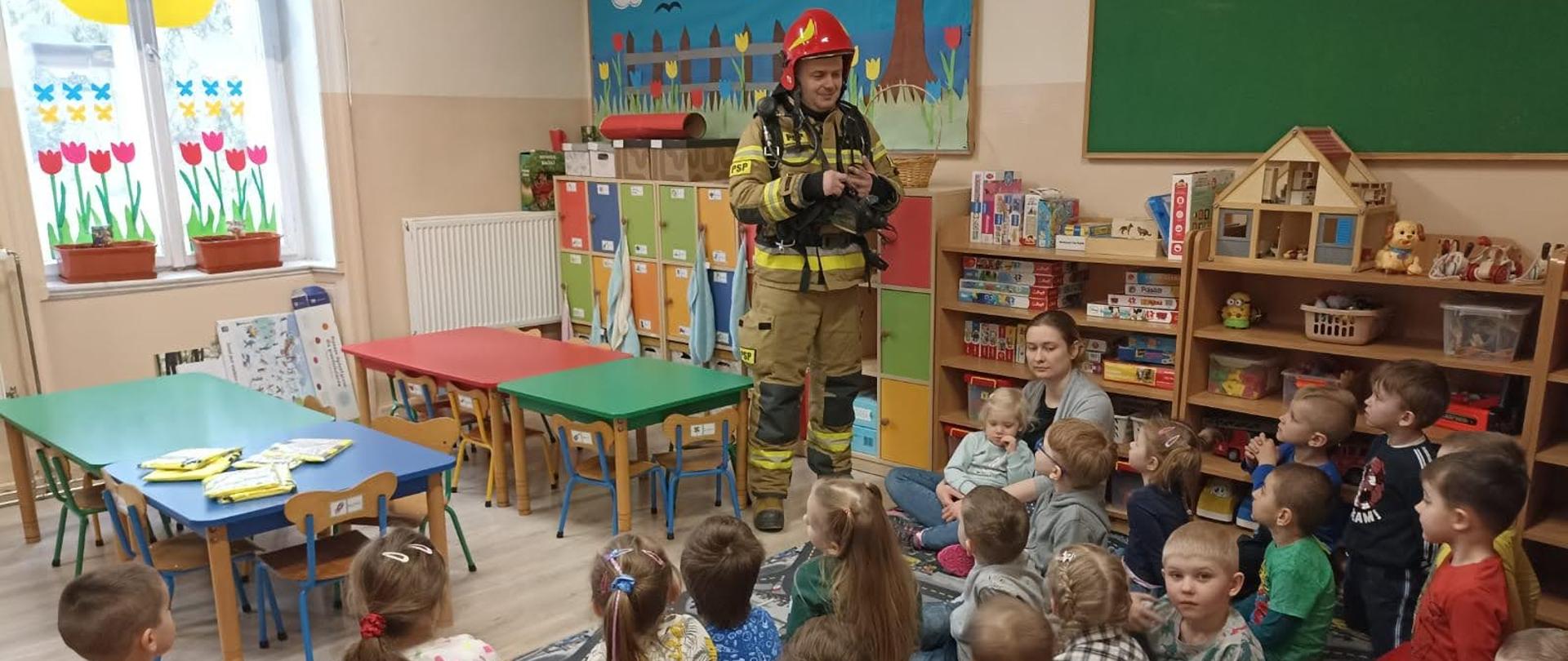 Na zdjęciu asp. sztabowy Marcin Ziółkowski podczas pogadanki w przedszkolu w Trzebczu Szlacheckim. Dzieci znajdują się w sali dydaktycznej. Funkcjonariusz prezentuje ubranie specjalne strażaka.