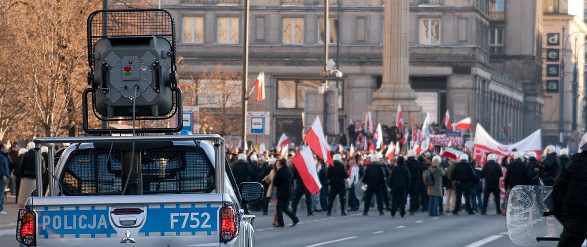 Zdjęcie przedstawia radiowóz oraz osoby z flagami Polski chodzące po ulicy.