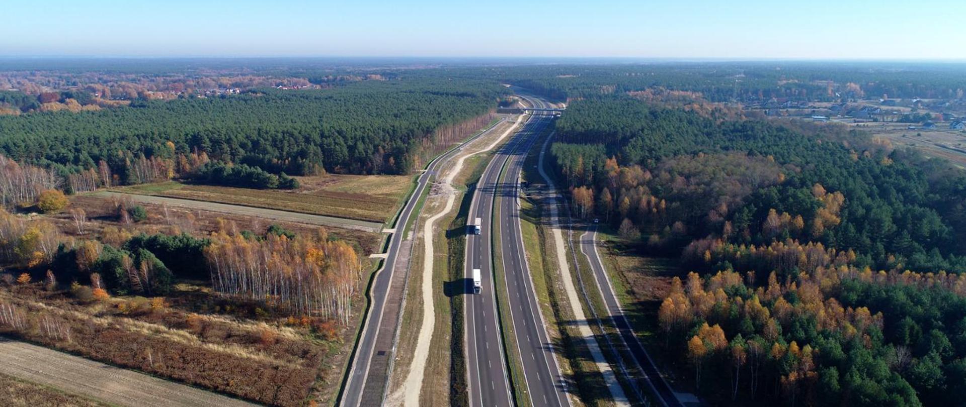 Widok z lotu ptaka na pojazdy jadące drogą ekspresową. Po obu stronach drogi las, w tle widoczny wiadukt.