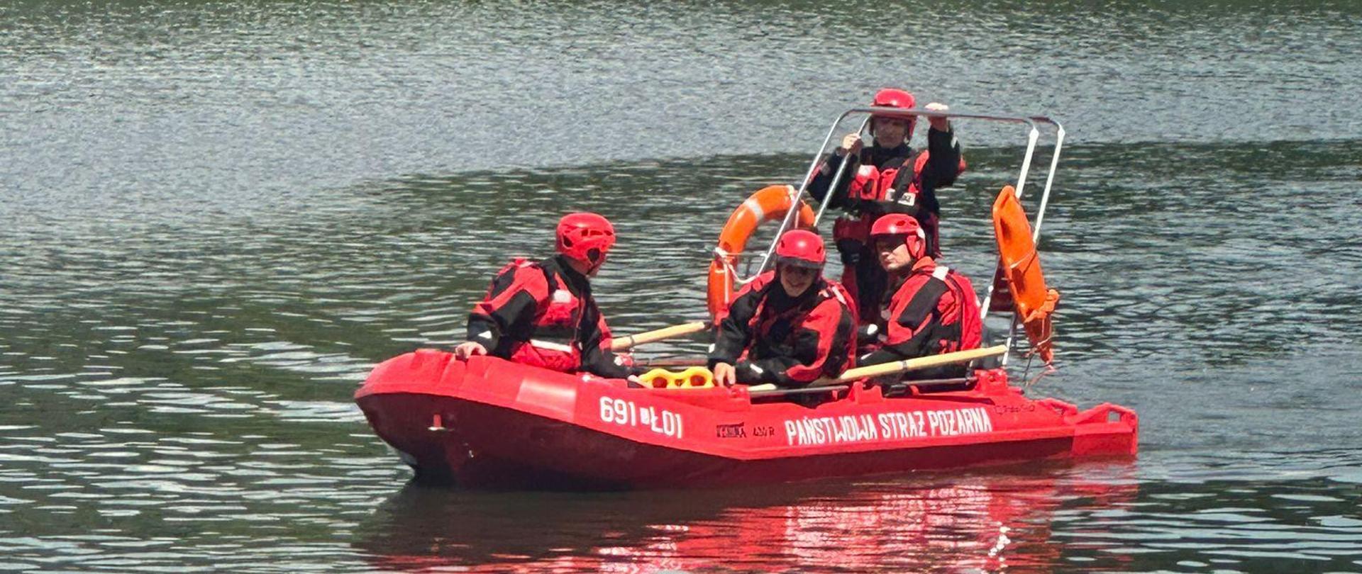 Czterech strażaków w łodzi ratowniczej w czerwonych skafandrach płynie po wodzie. Za nimi w tle zarośla i drzewa liściaste. 