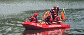 Czterech strażaków w łodzi ratowniczej w czerwonych skafandrach płynie po wodzie. Za nimi w tle zarośla i drzewa liściaste. 