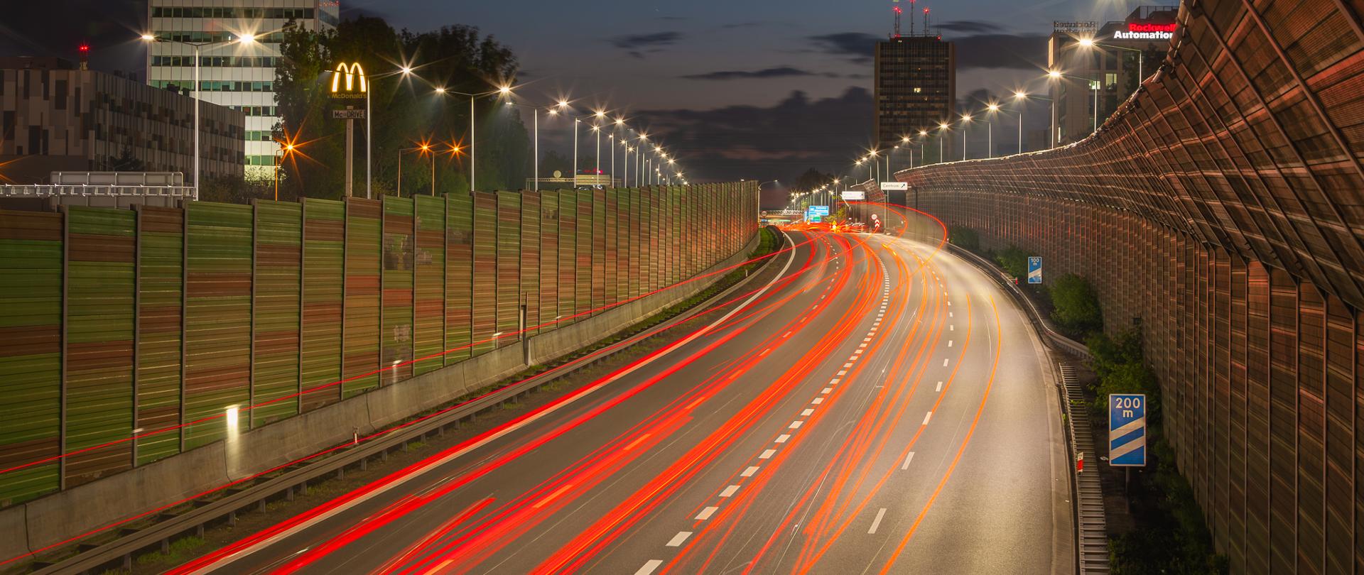 Na nocnym zdjęciu widzimy autostradę z efektownymi śladami świetlnymi pojazdów, co wskazuje na wykorzystanie długiego czasu naświetlania w fotografii. Autostrada jest otoczona ekranami dźwiękochłonnymi po obu stronach, a latarnie uliczne oświetlają drogę. W tle widoczne są wysokie budynki z podświetlonymi szyldami. Niebo jest ciemne, pokryte chmurami, a cała scena oddaje dynamikę i ruch na autostradzie nocą.