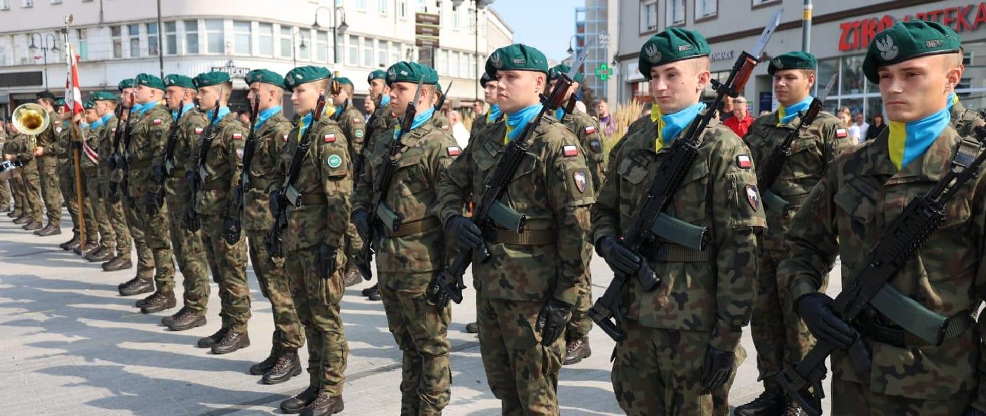 Żołnierze stoją w szeregu. Ubrani są w mundury. Każdy z nich trzyma broń. 