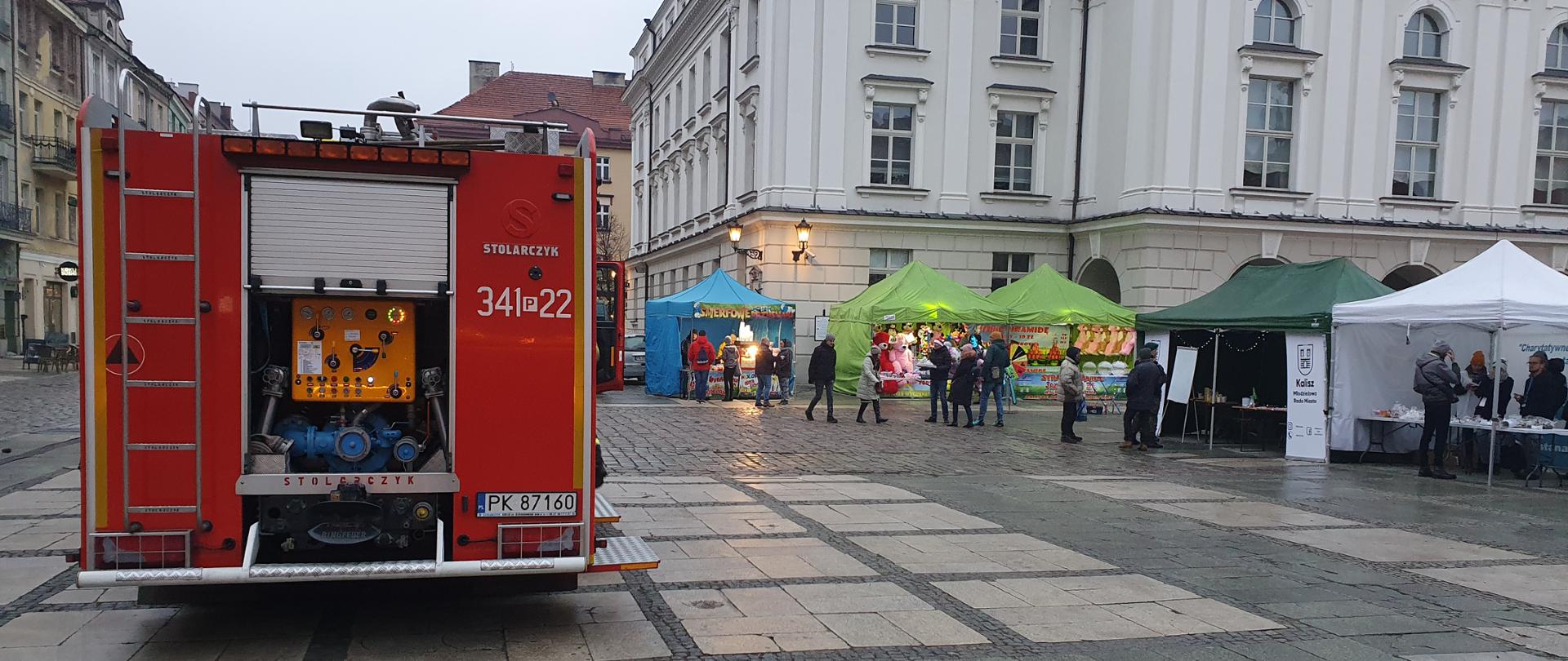 Na zdjęciu widać samochód gaśniczy należący do JRG 1 Kalisz. Samochód ma kolor czerwony, ma oznakowanie oraz lampy ostrzegawcze kolory niebieskiego. Skrytki zabudowy są otwarte. Pojazd stoi na płycie rynku. W tle widać zabudowania Głównego Rynku oraz namioty wystawowe.