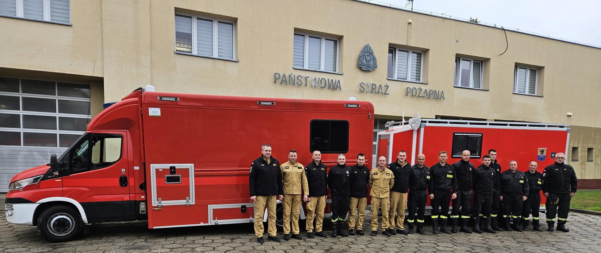 Na tle remizy PS stoi zaparkowany samochód SDŁ wraz z przyczepą. Przed samochodem w rzędzie strażacy uczestniczący w działaniach.