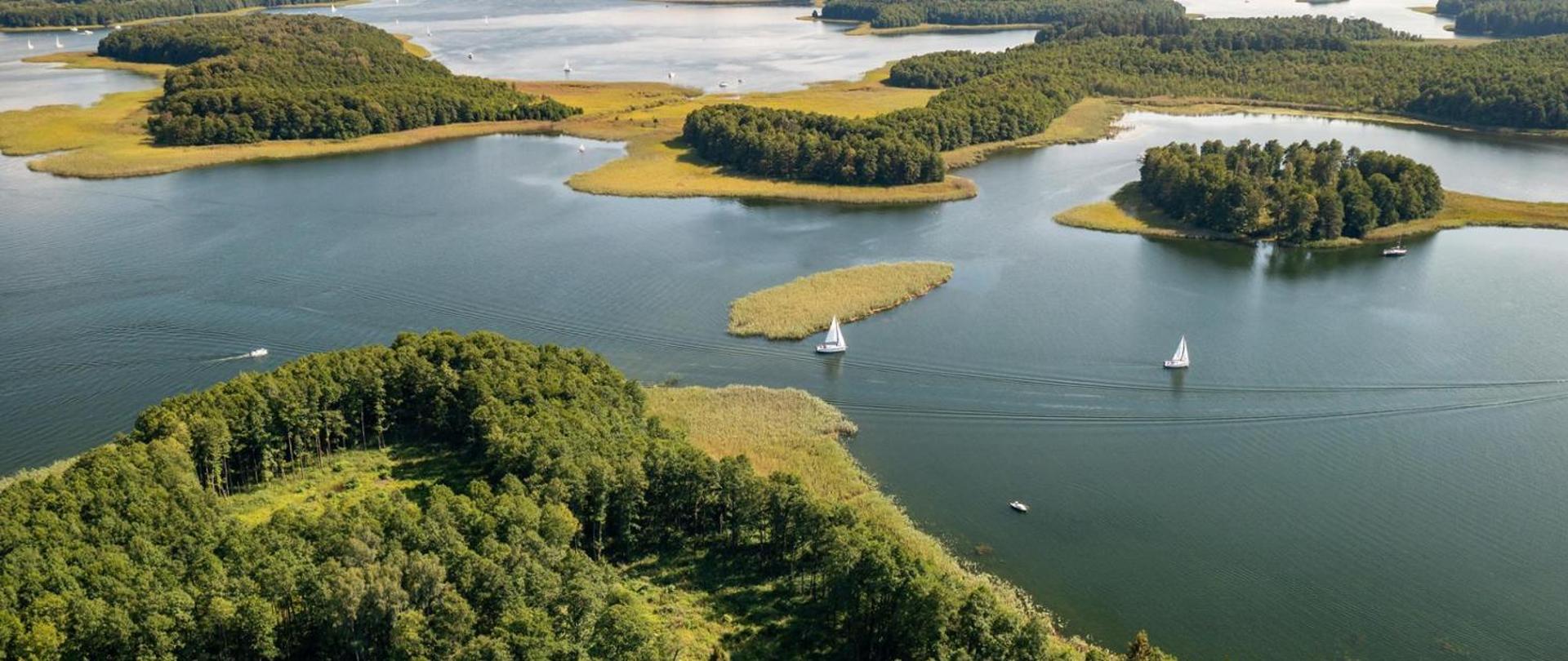 Panorama. Widok na jezioro i porośnięte lasem wyspy na jeziorze. Po jeziorze pływają białe żaglówki.