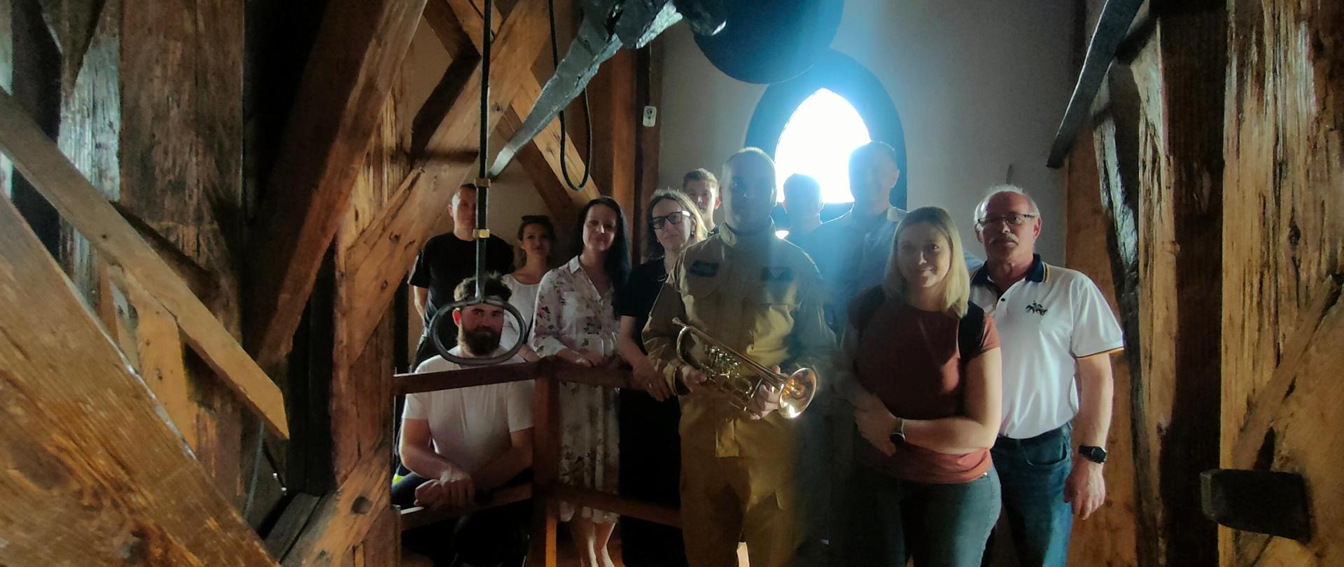Pomieszczenie na szczycie wieży Kościoła Mariackiego. Zdjęcie pamiątkowe uczestników z trębaczem. Pomiędzy uczestnikami ubrany w umundurowanie służbowe strażak trzymający w ręku trąbkę koloru złotego. 
