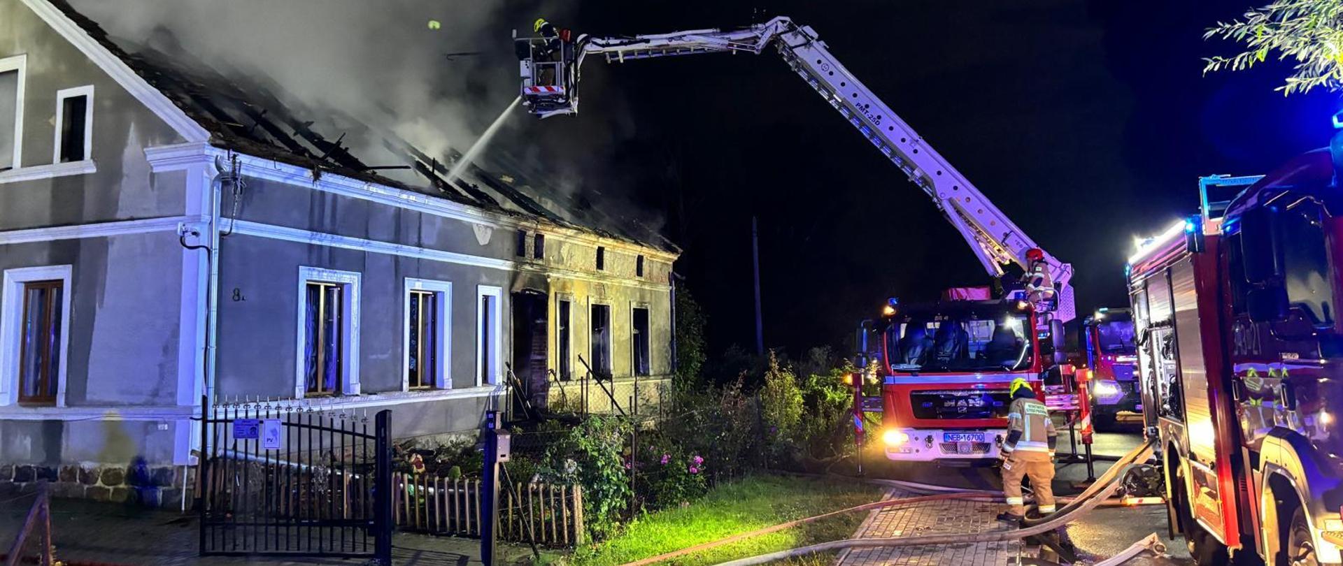 Strażacy przy użyciu podnośnika podają prąd wody na dach domu wielorodzinnego. Widać kłęby dymu wydostające się ze spalonego dachu. Obok budynku widoczne są pojazdy pożarnicze i sylwetki strażaków. Wszystko dzieje się w nocnej scenerii. 