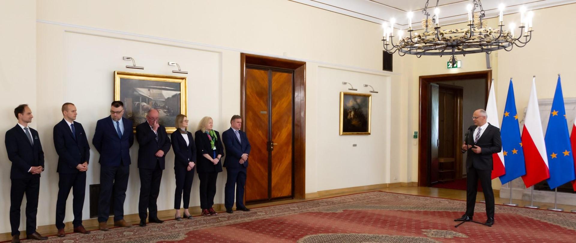 Grupa kobiet i mężczyzn stoi przy ścianie, niedaleko nich mężczyzna przemawia do mikrofonu, a za nim stoją flagi Polski i Unii Europejskiej 