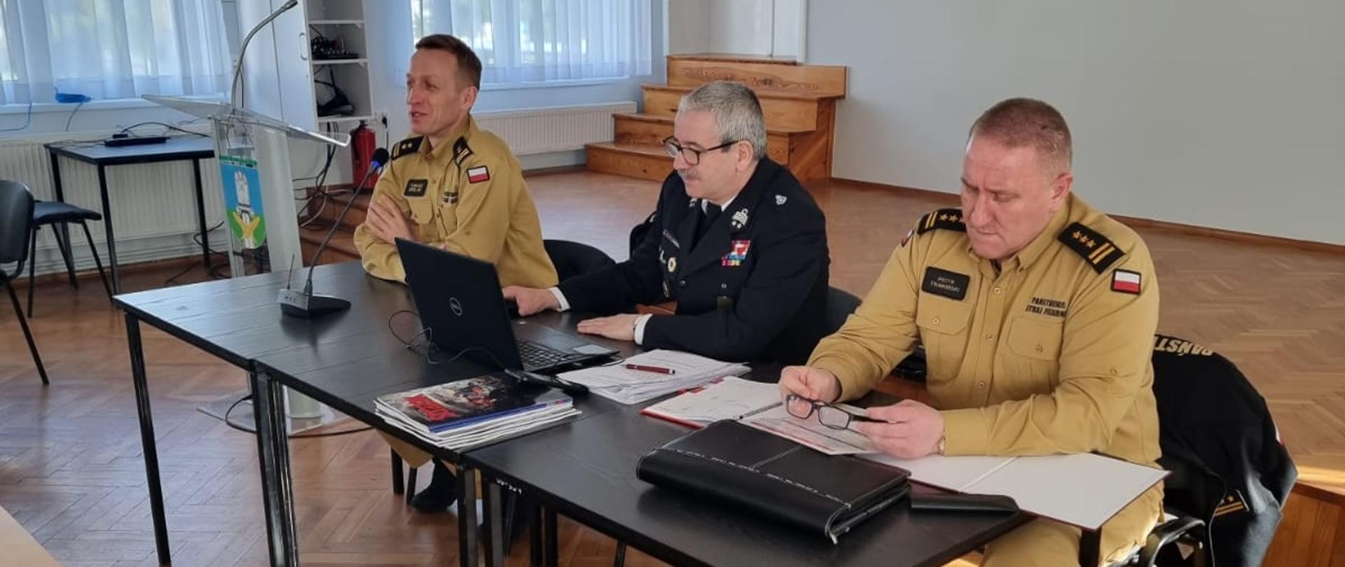 Zdjęcie przedstawia trzech strażaków siedzących przy stole prezydialnym, za nimi godło Polski i godło Kołaczkowa