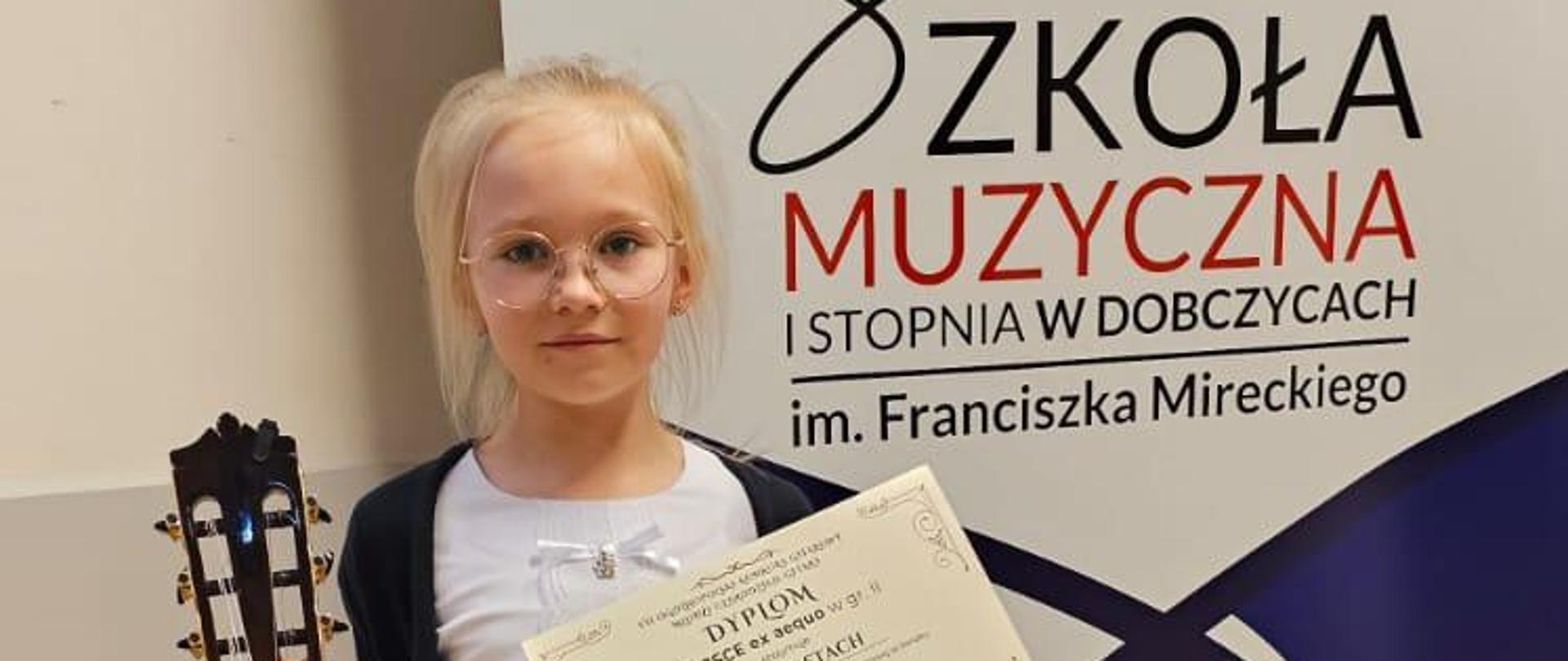 Zuzanna Stach w VII Ogólnopolskim Konkursie Gitarowym „Młodzi czarodzieje gitary” w Dobczycach. Zdjęcie uczennicy z gitarą, w tle rollup reklamujący konkurs.