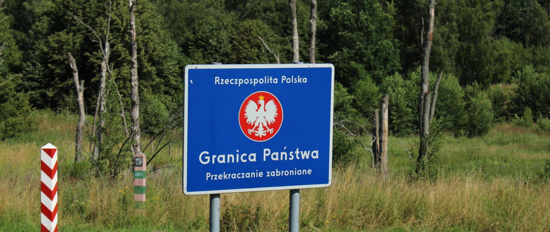 Zdjęcie przedstawia tablicę informującą o granicy państwa. W tle znajduje się las i słupki graniczne.