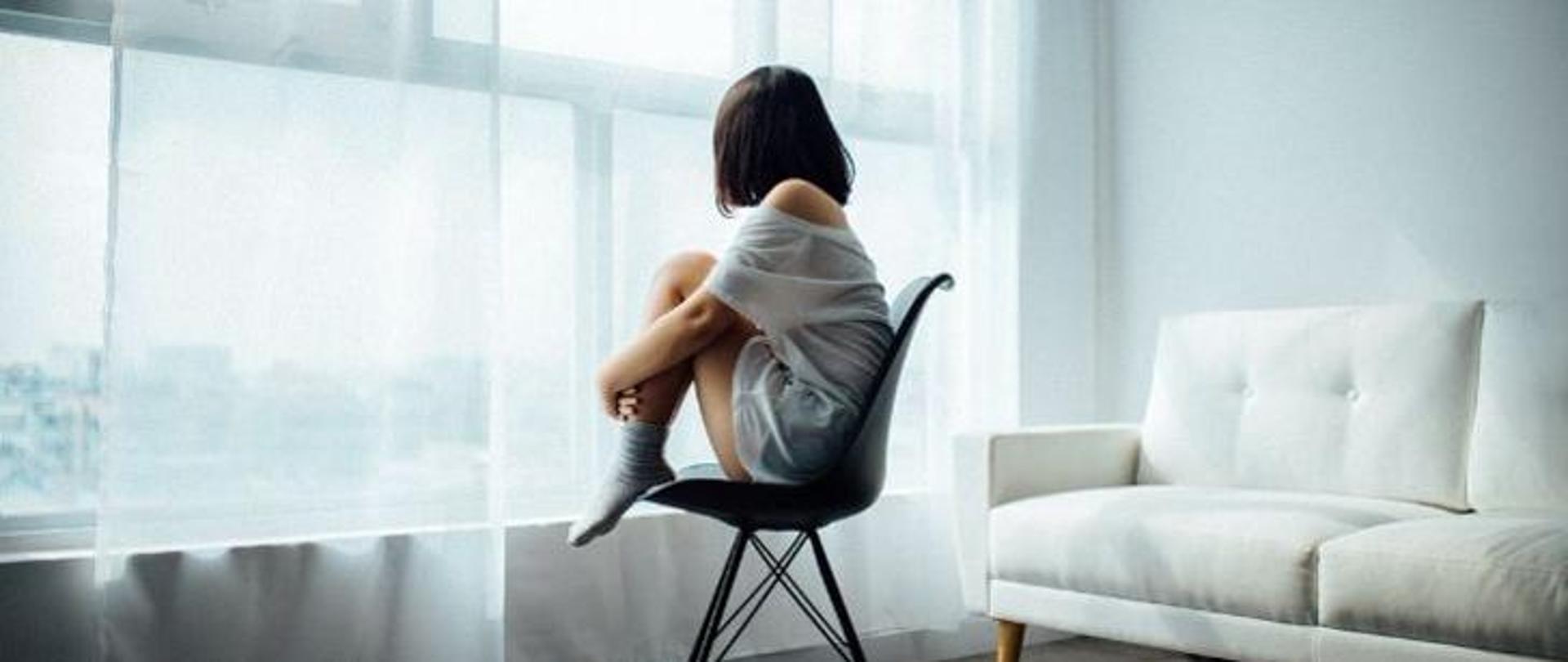 Dziewczyna w czarnych włosach siedząca na krześle z przykurczonymi nogami i zapatrzona w okno