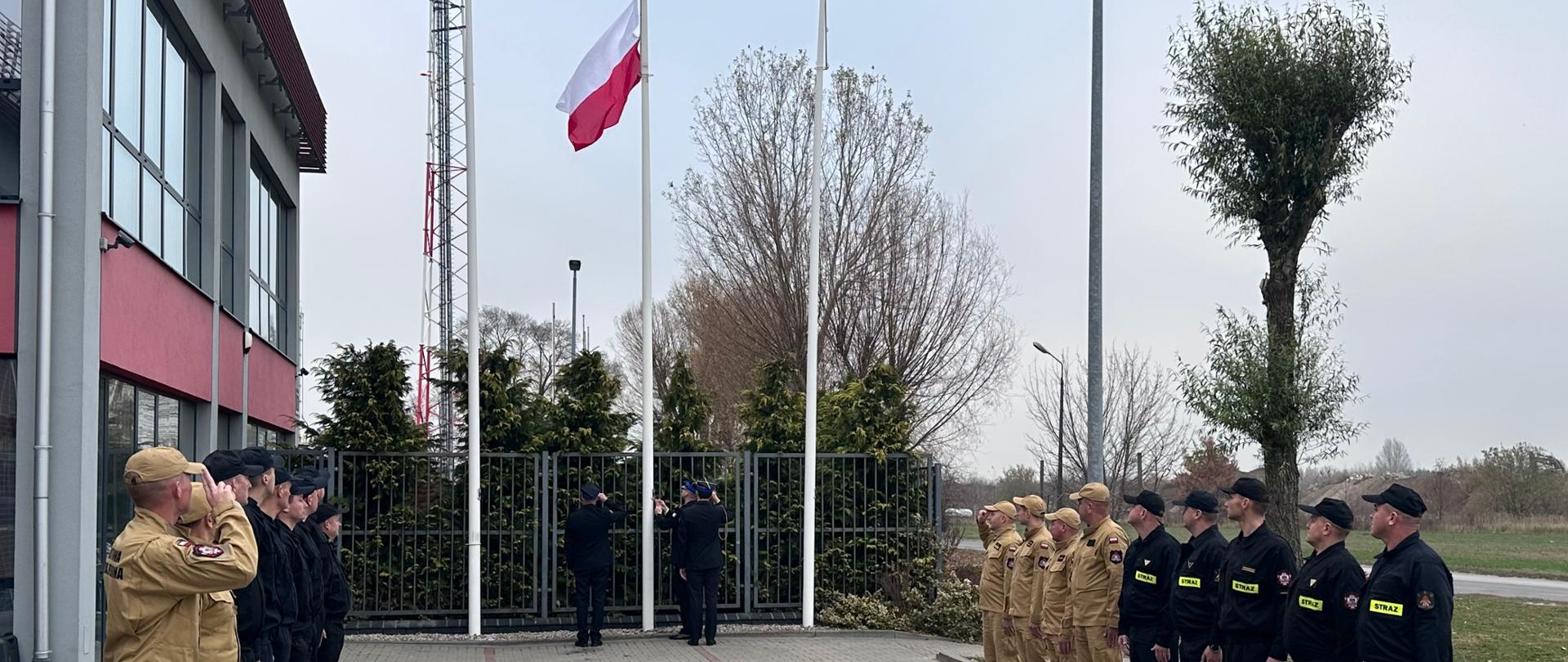 Na zdjęciu widać strażaków Państwowej Straży Pożarnej w Radomiu, podczas uroczystej zmiany służby w dniu 11 listopada 2024 roku. W czasie zmiany służby, z okazji 106 rocznicy Odzyskania Niepodległości przez Polskę nastąpiło podniesienie flagi państwowej. Strażacy w momencie podnoszenie flagi państwowej, zwracają głowę w stronę flagi, a dowódcy zmian i dowódca jednostki oddają honor. 