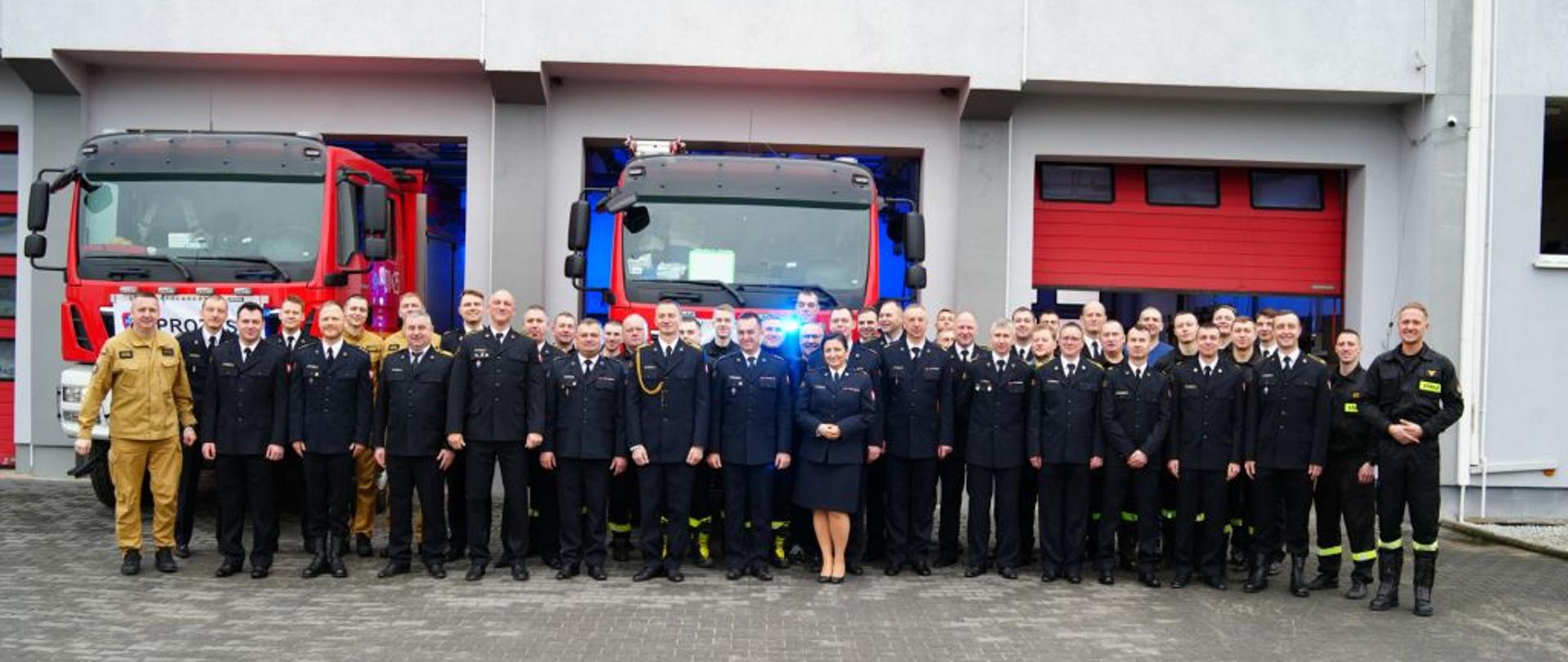 Zdjęcie wykonane przed budynkiem straży pożarnej. Przed remizą stoi duża grupa strażaków w mundurach galowych i bojowych. W tle widać dwa czerwone wozy strażackie, a budynek remizy jest nowoczesny i dobrze utrzymany. Strażacy stoją w równym rzędzie, patrząc w stronę aparatu. Ujęcie podkreśla jedność i gotowość strażaków do działania.