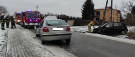 Zdjęcie przedstawia uszkodzony pojazd, z lewej za nim stoją samochody strażackie i strażacy w ubraniach bojowych. Po prawej na poboczu stoi drugi uszkodzony pojazd a przy nim strażak odłącza akumulator.