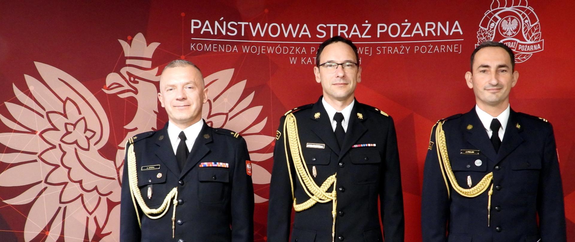 Na tle czerwonej ścianki z białym orłem w koronie po lewej stronie a po prawej logo Państwowej Straży Pożarnej. Na środku napis Państwowa Straż Pożarna. Komenda Wojewódzka PSP w Katowicach, stoją w mundurach wyjściowych, w środku śląski komendant wojewódzki nadbryg. Wojciech Kruczek, po prawej nowo powołany Komendant Powiatowy PSP w Żywcu - mł. bryg. Jarosław Polak, po lewej Zastępca Komendanta Miejskiego PSP w Piekarach Śląskich - mł. bryg. Arkadiusz Spera
