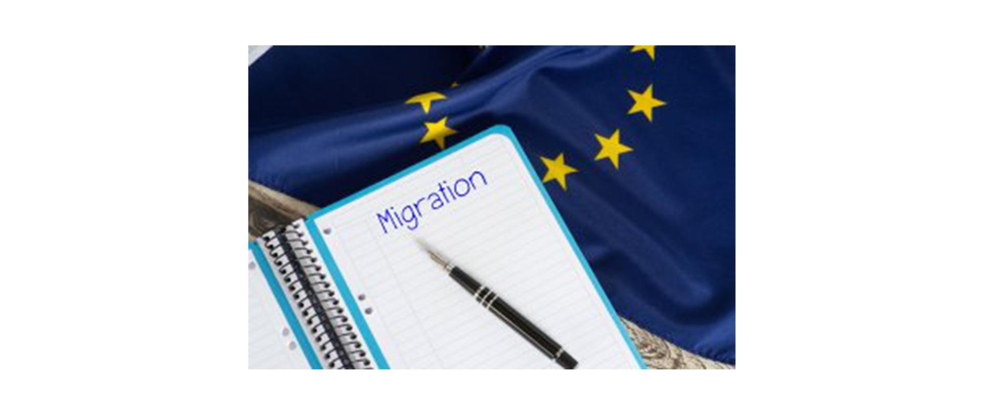 Na zdjęciu: notatnik z napisem "Migration"; leży na nim długopis. W tle flaga UE.