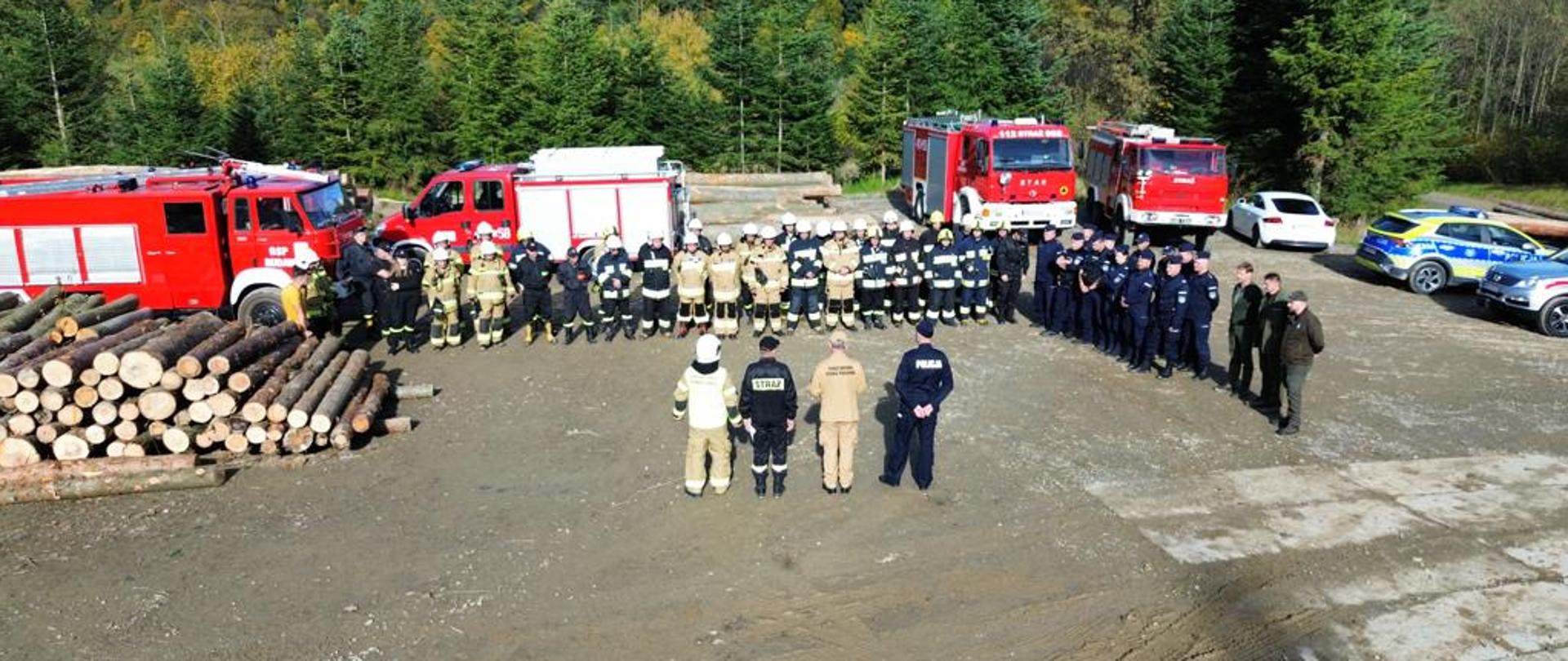 Kolorowa fotografia wykonana na zewnątrz w pogodny dzień. Przedstawia strażaków, policjantów oraz służbę leśną na zbiórce przed ćwiczeniami. W tle samochody pożarnicze, policji oraz ścięte i składowane drewno.