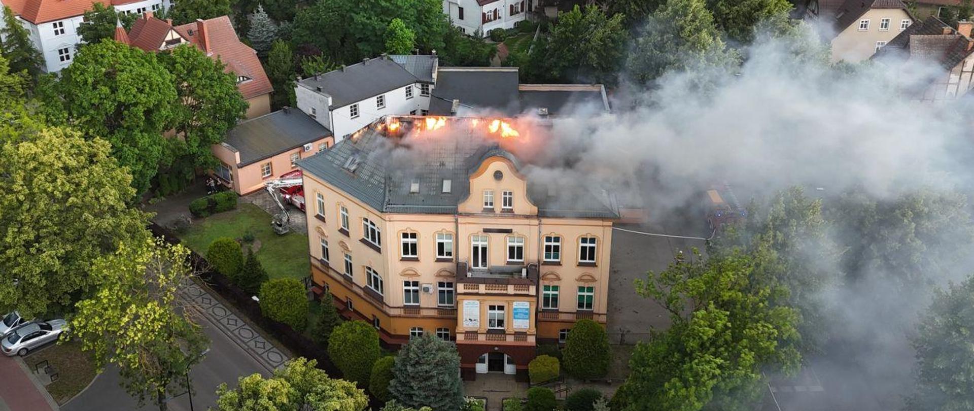 Zdjęcie wykonane z drona. Centralny punkt zdjęcia stanowi budynek. Wysoka kamienica, z dachu wydostaje się ogień.