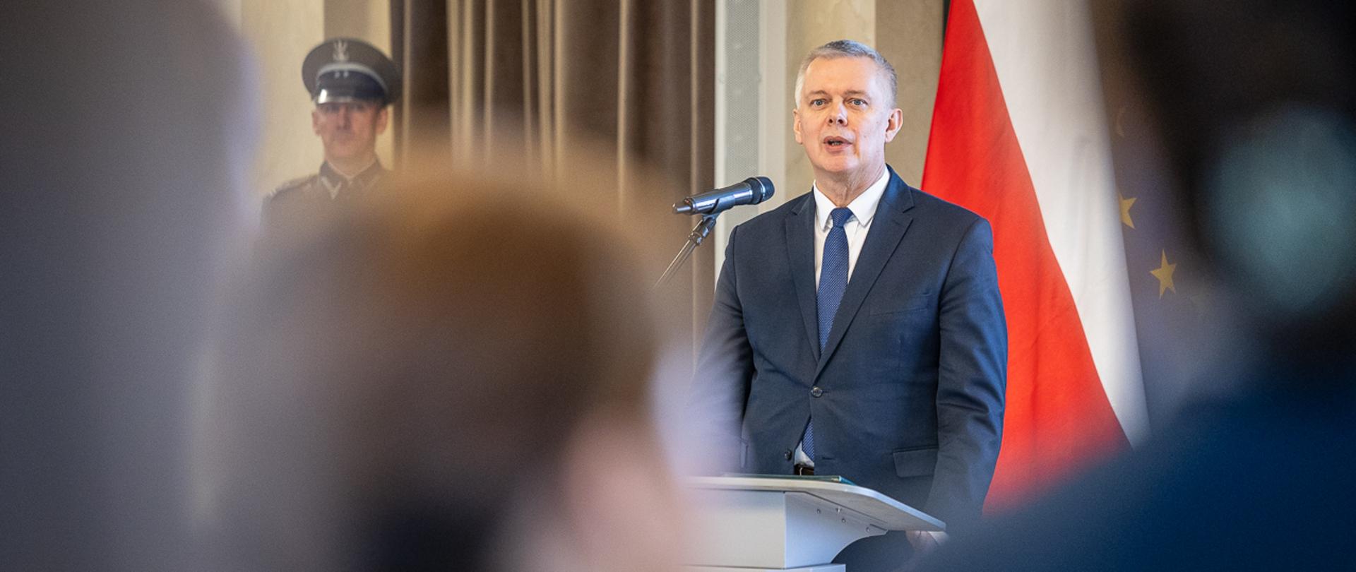 Mężczyzna za mównicą na tle flag Polski i UE przemawia do zebranych, po lewej w nieostrości mężczyzna w mundurze Korpusu Ochrony Pogranicza. 