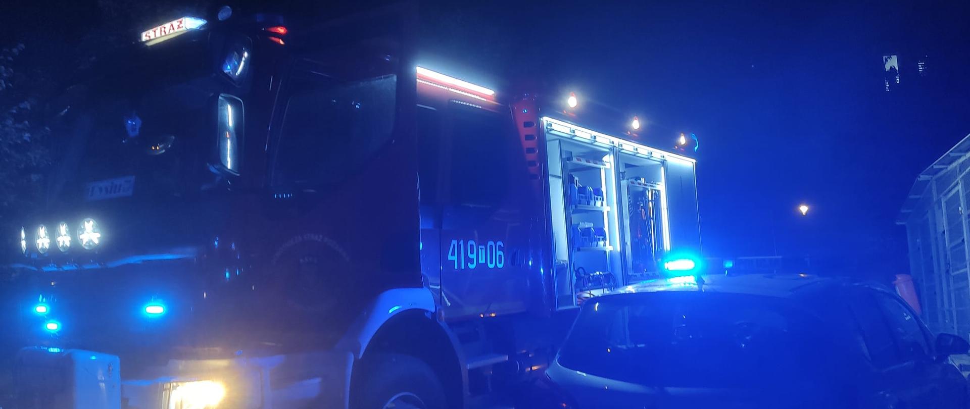Na zdjęciu widoczny samochód ratowniczo-gaśniczy oraz radiowóz policyjny. Pojazdy z włączoną sygnalizacją świetlną w kolorze niebieskim. Zdjęcie zrobione w porze nocnej. 
