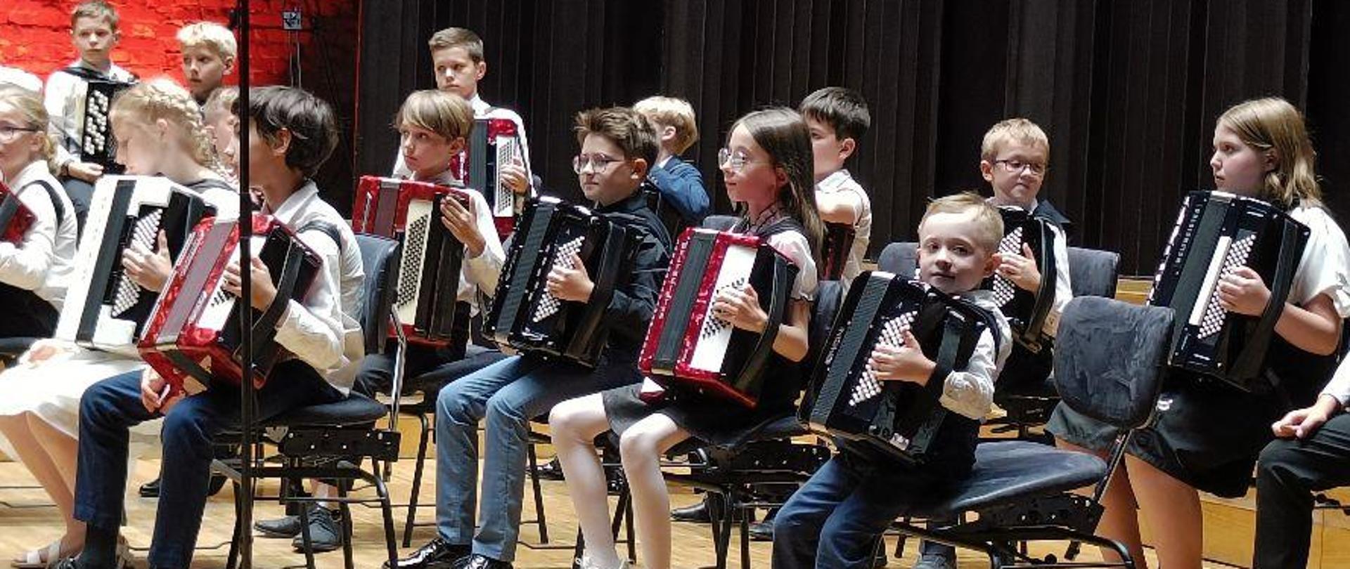 Uczniowie trzymający akordeony, siedzący na scenie Filharmonii. Na dole fotografii widoczna publiczność.